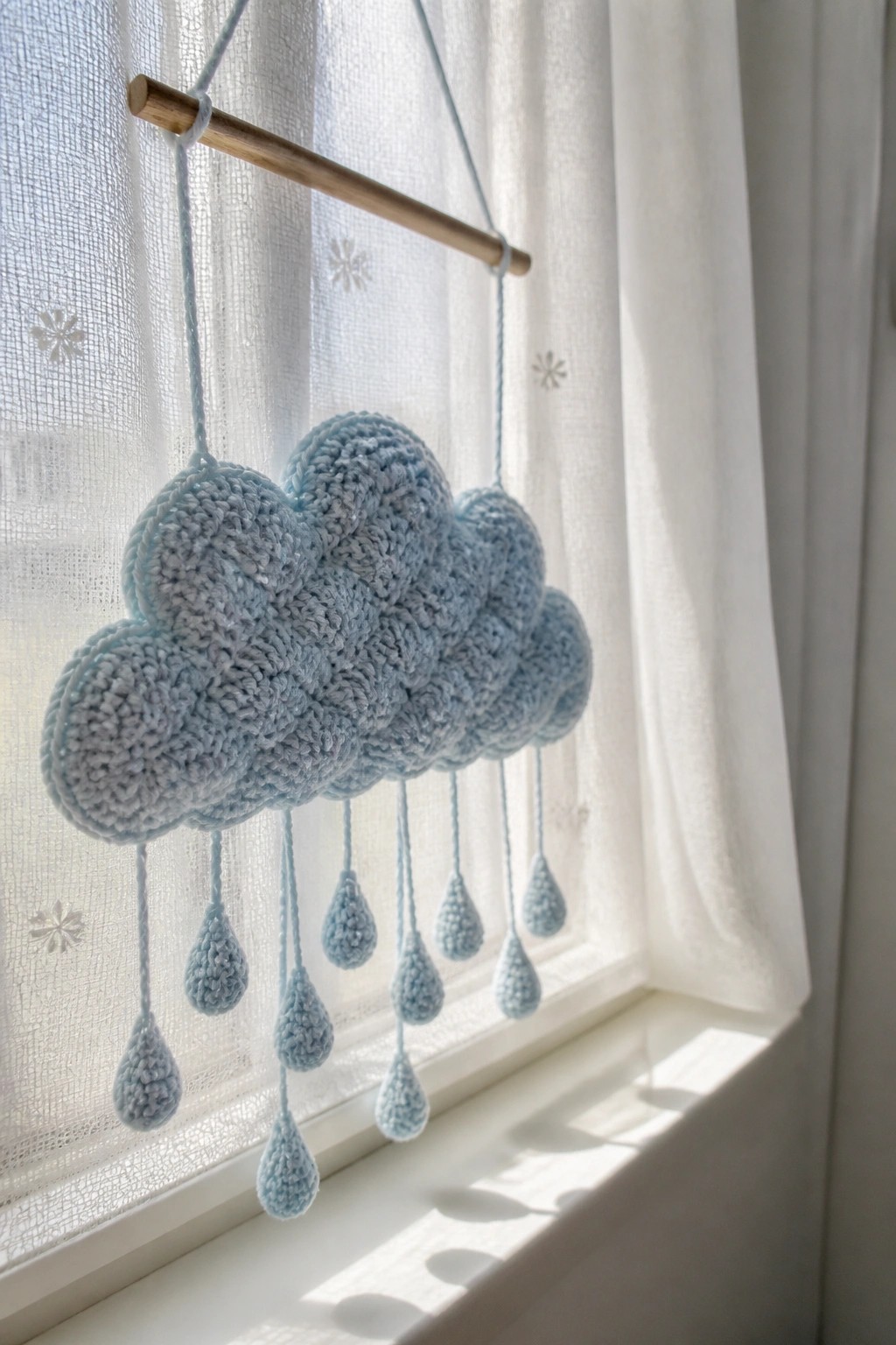A light blue crocheted cloud with dangling teardrop shapes hangs from a wooden dowel in front of a window.
