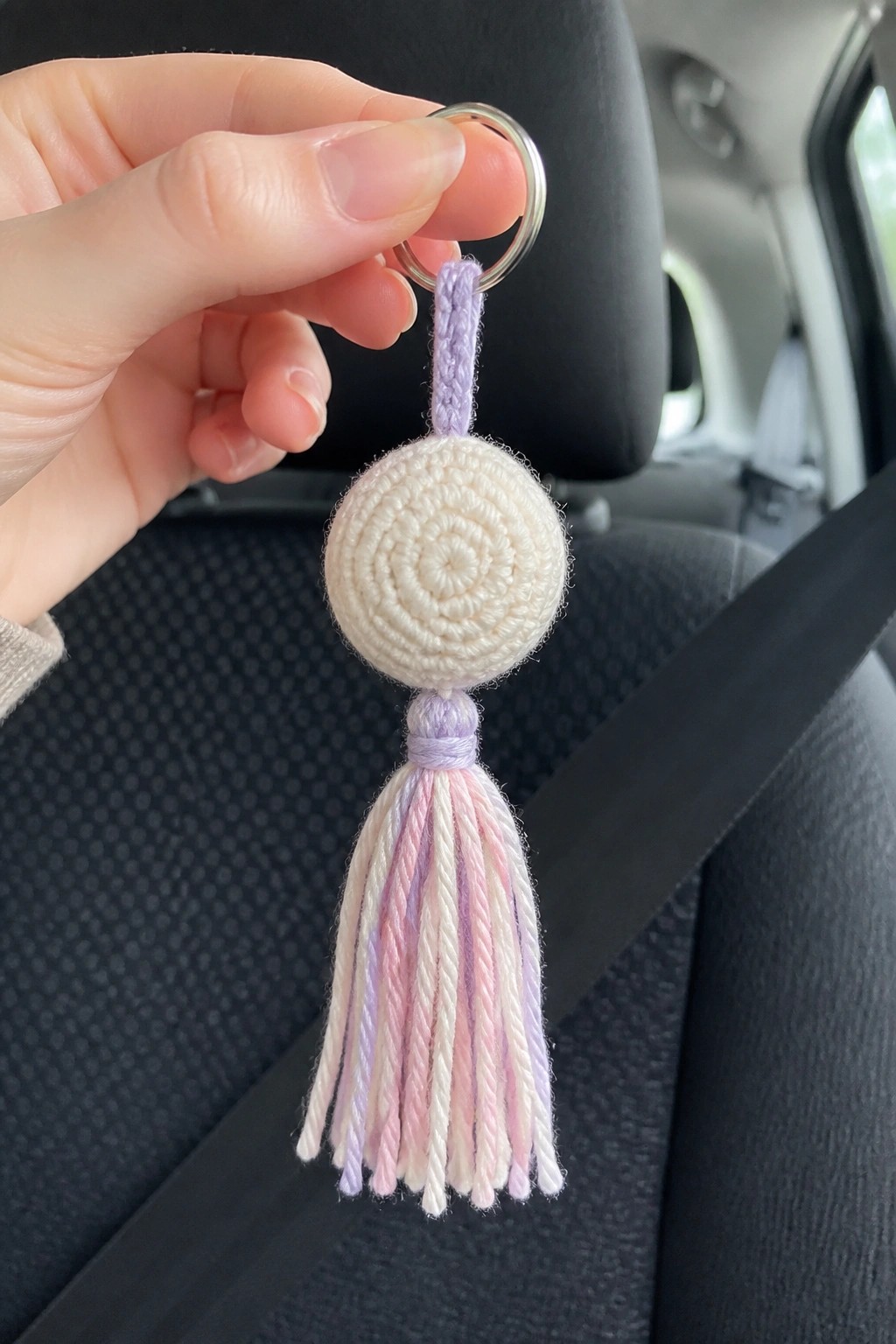 Hand holds crocheted white keychain with lavender cord and pastel yarn tassel in car.