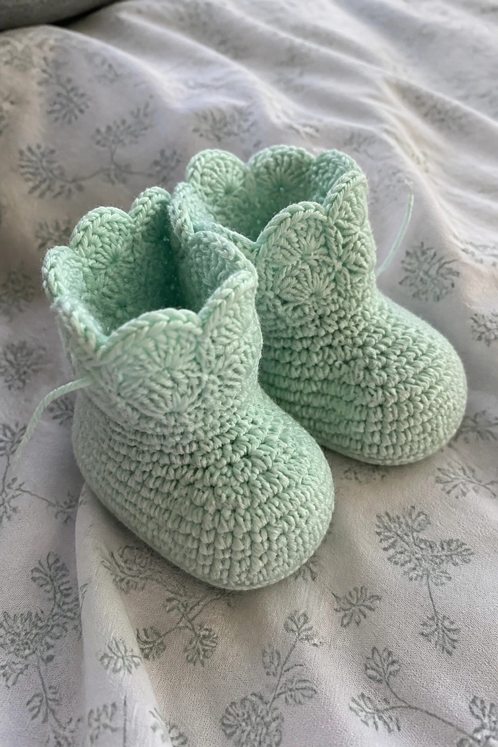 Mint green crocheted baby booties with scalloped tops on floral fabric