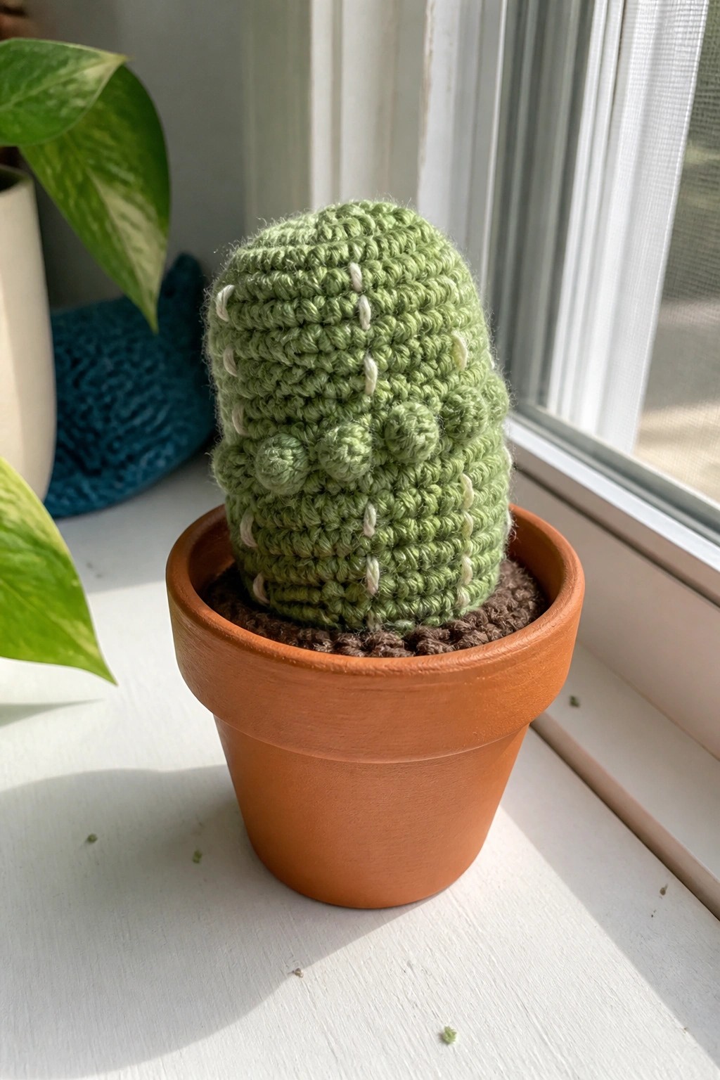 A green crocheted cactus with white spine details sits in a small terracotta pot on a white windowsill.