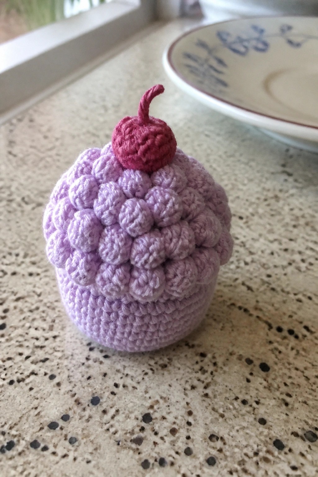 Crocheted purple cupcake with bumpy top and red cherry on speckled countertop.