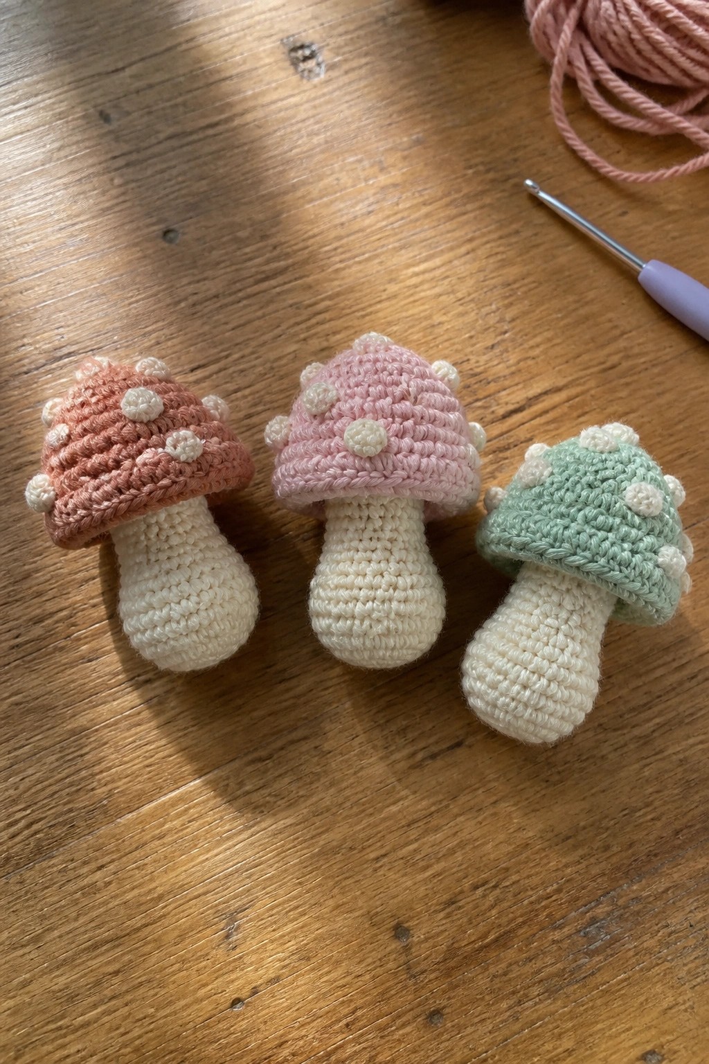 Three crocheted mushrooms in peach, pink, and mint green on wood
