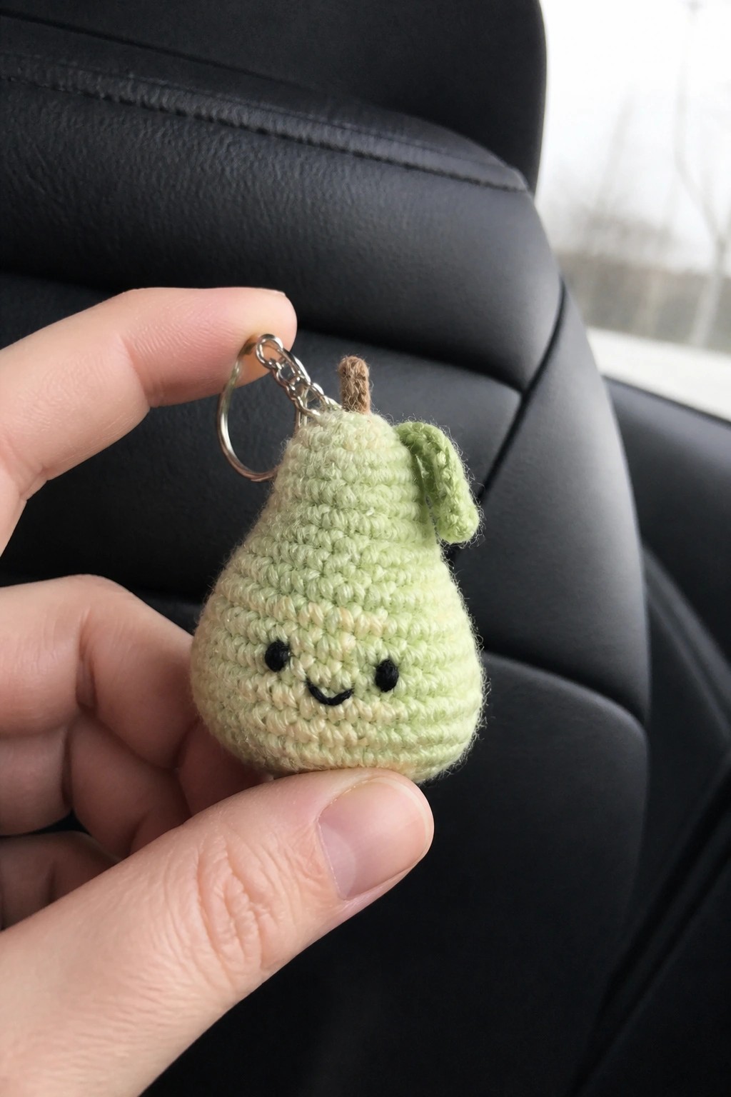 Hand holding light green crocheted pear keychain with smiling face and metal ring.