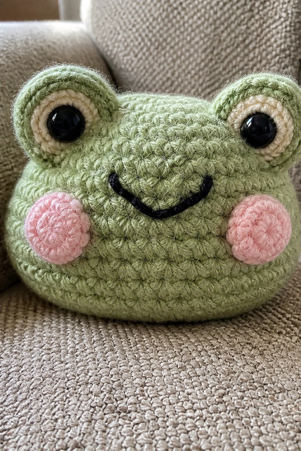 Green crocheted frog with pink cheeks, large black eyes, and smiling mouth on fabric