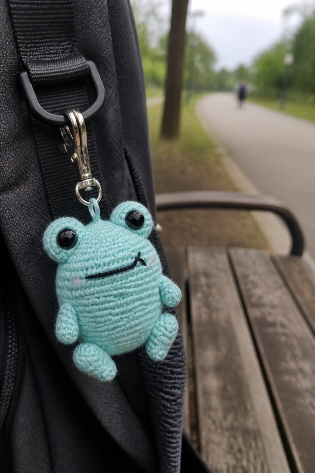 Crocheted turquoise frog keychain clipped to black bag on wooden park bench