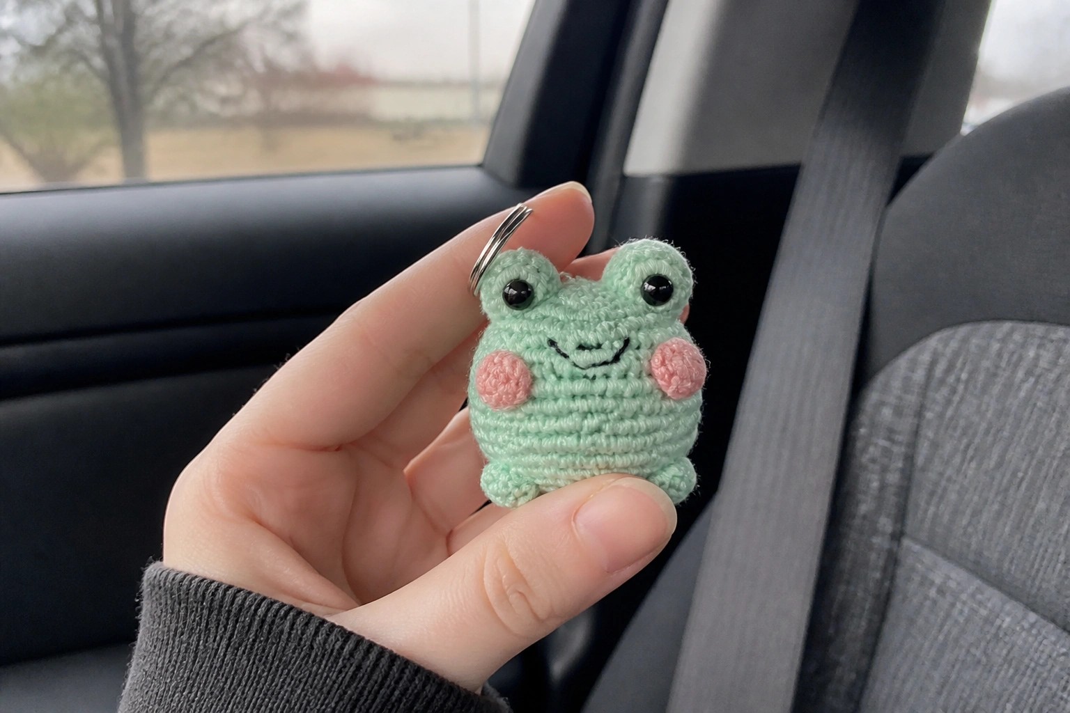 Hand holds mint green crocheted frog keychain with pink cheeks in car.