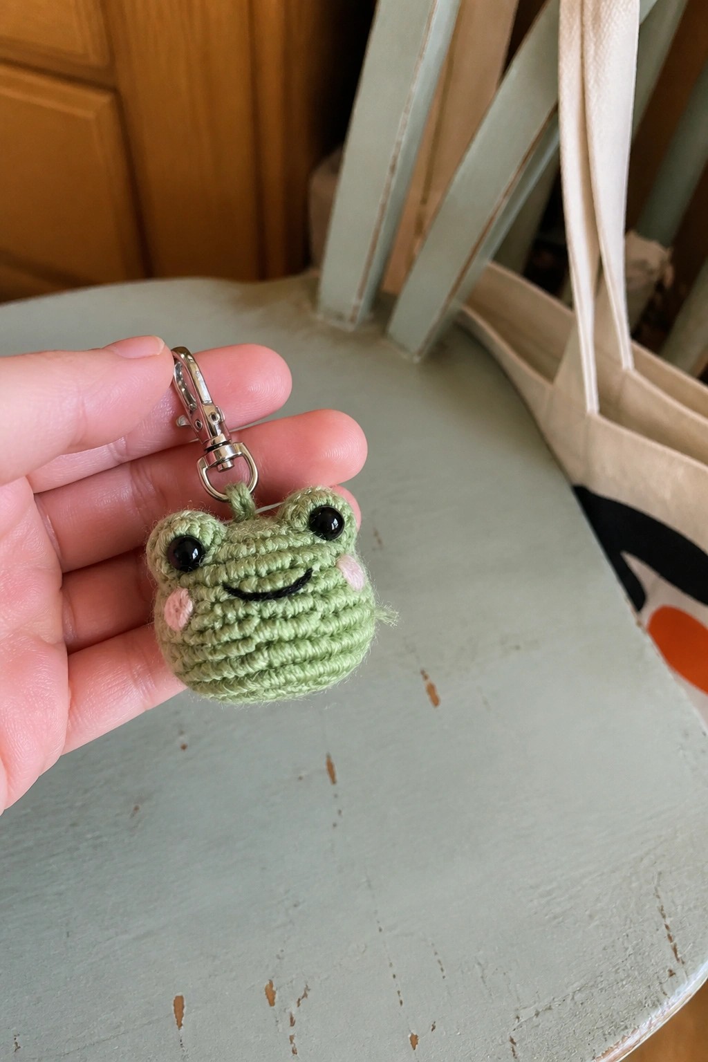 Hand holding green crocheted frog keychain with pink cheeks, black eyes, and metal clip