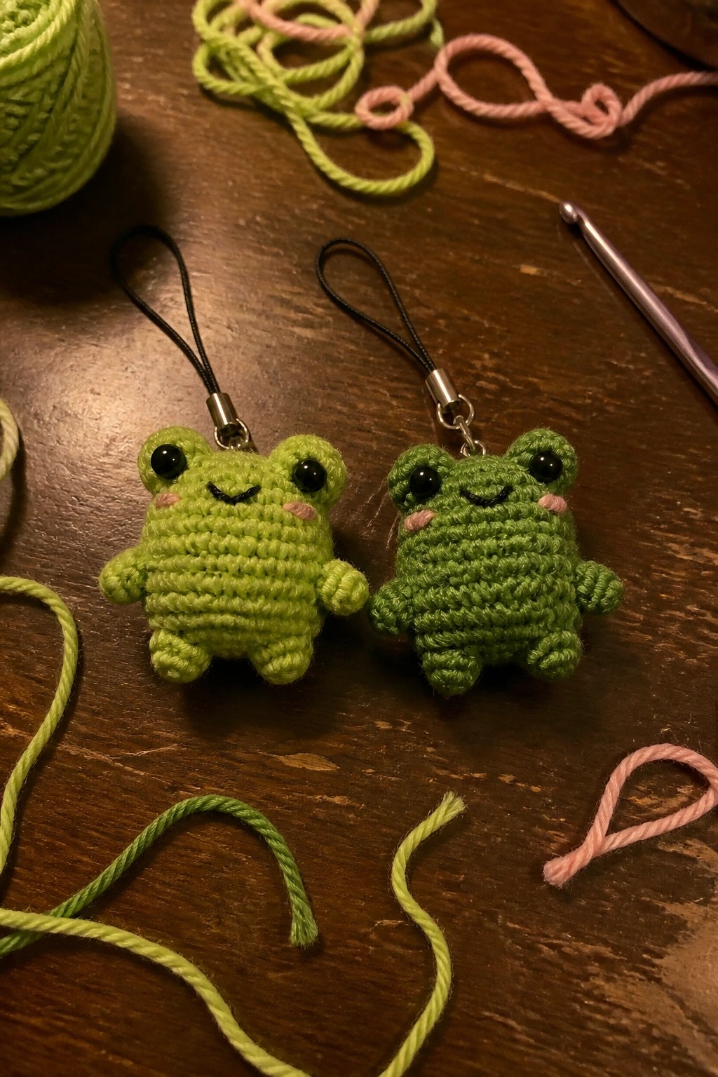 Two green crocheted frog keychains with pink cheeks on wooden table amid yarn