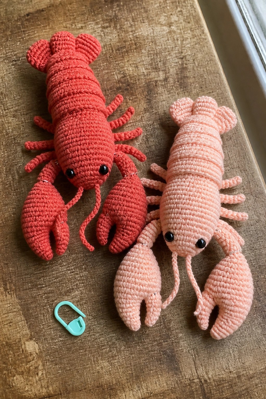 Two crocheted lobsters, red and pink, on wooden table with turquoise marker