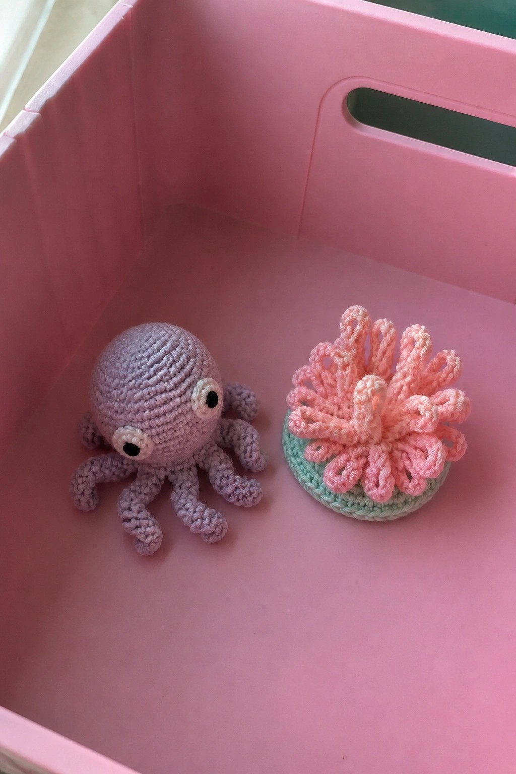 A crocheted purple octopus and a pink crocheted sea anemone sitting side by side in a pink plastic bin.