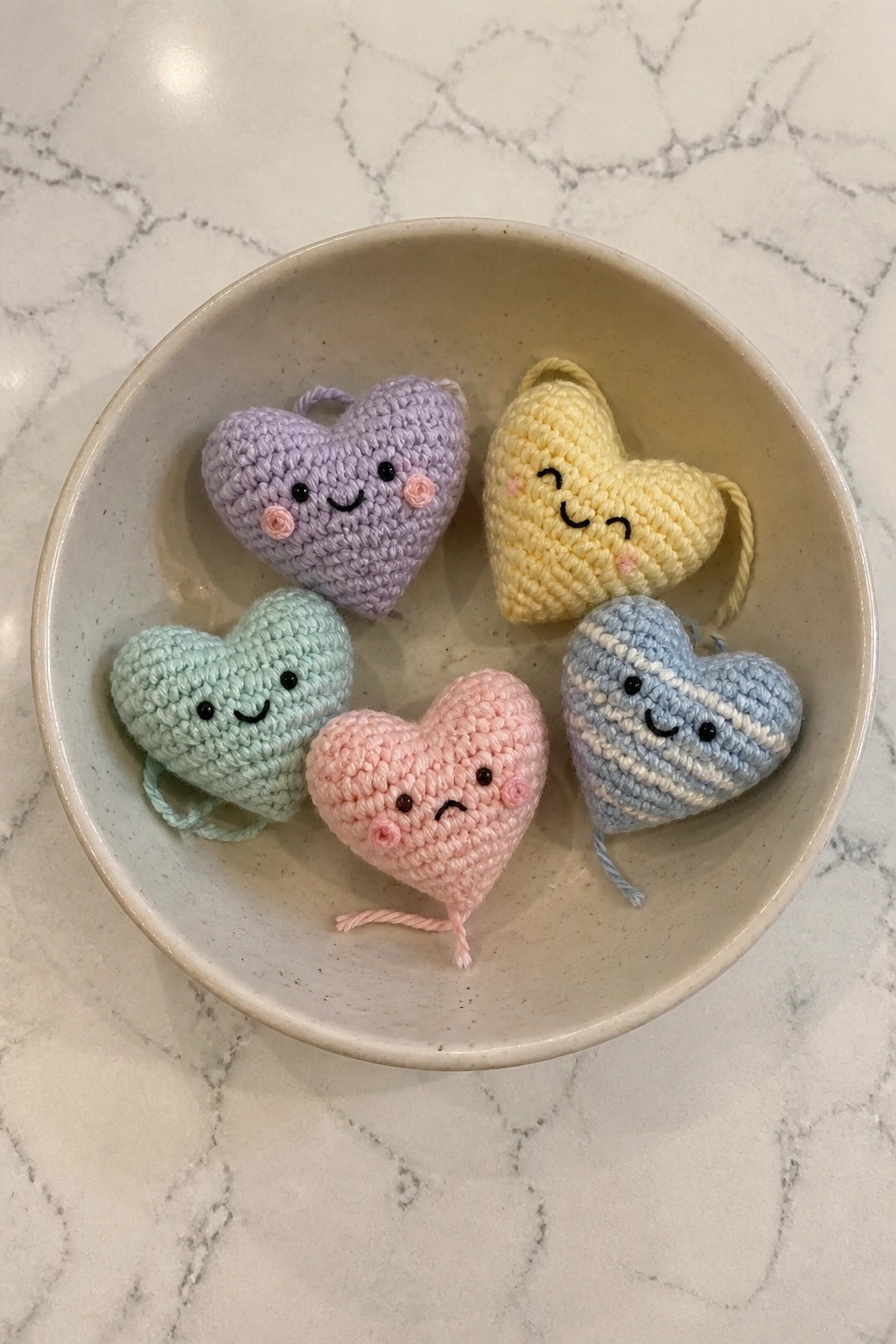Five pastel crocheted hearts with faces in a beige ceramic bowl on marble.