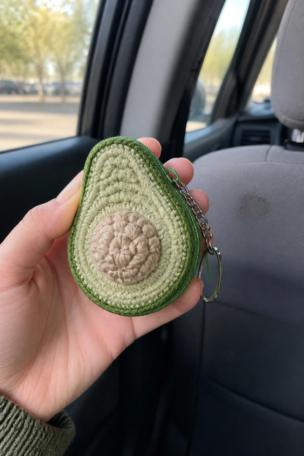 Hand holding crocheted avocado keychain inside car with gray seats visible.