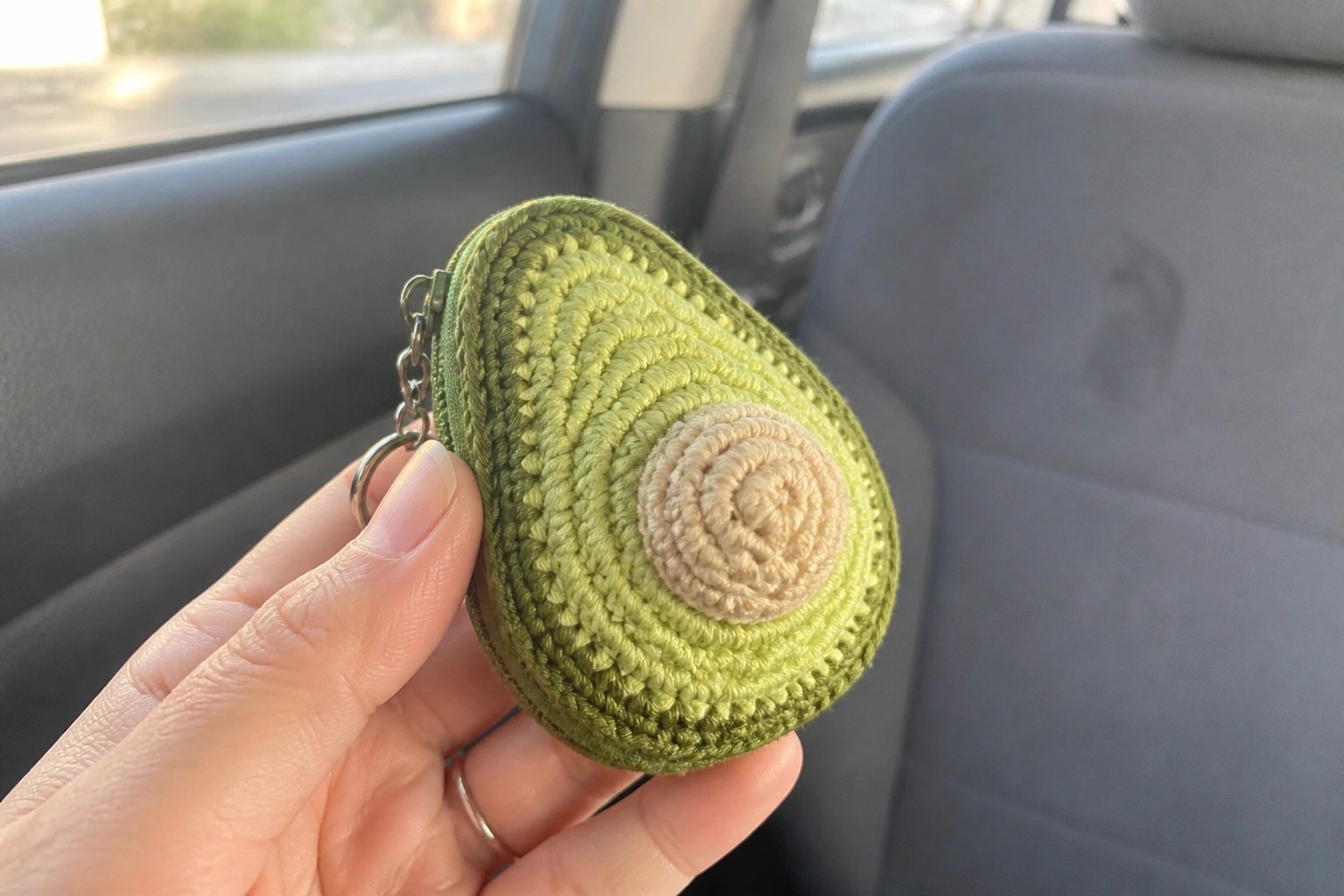 Hand holding crocheted avocado keychain inside car with gray seats visible.
