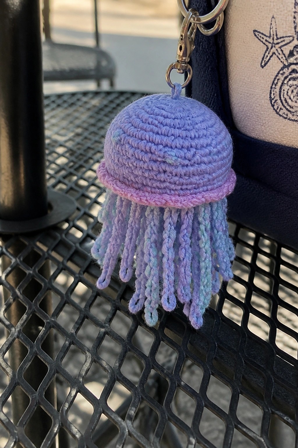 Purple crocheted jellyfish keychain with pastel tentacles beside starfish-printed bag on mesh table.