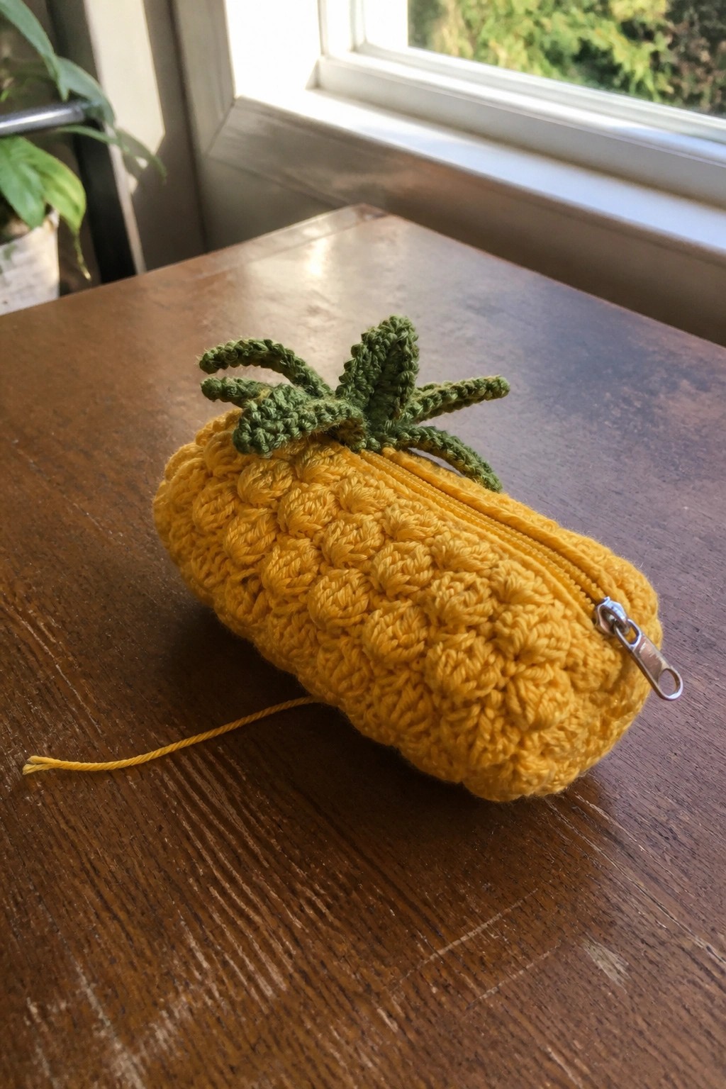 A yellow crocheted pineapple-shaped pouch with green leaves on top and a silver zipper along the side rests on a wooden table.
