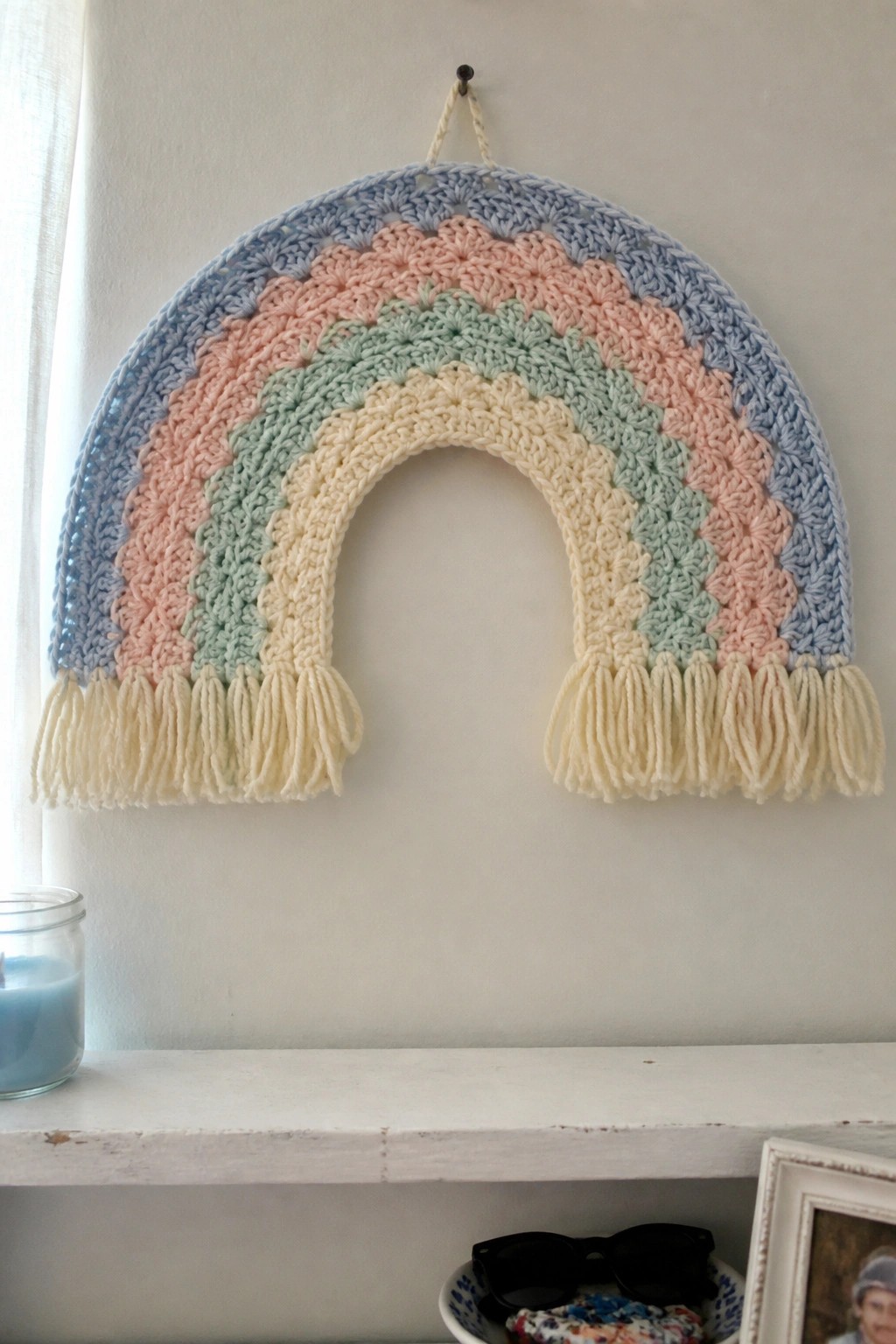 Pastel crocheted rainbow with yellow fringes hanging above white mantel shelf.
