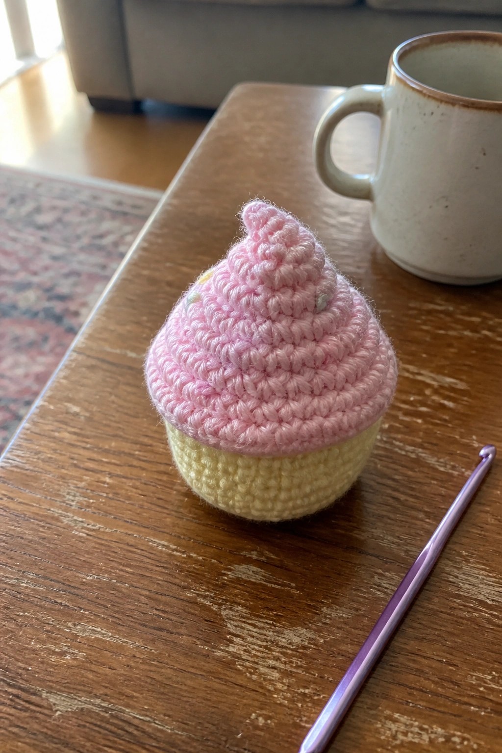Pink crocheted cupcake on wooden table beside purple hook and mug