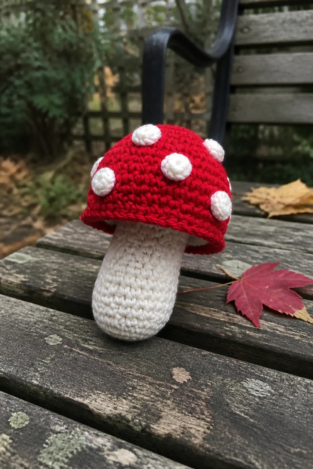 Crocheted red mushroom with white spots on wooden bench with autumn leaves