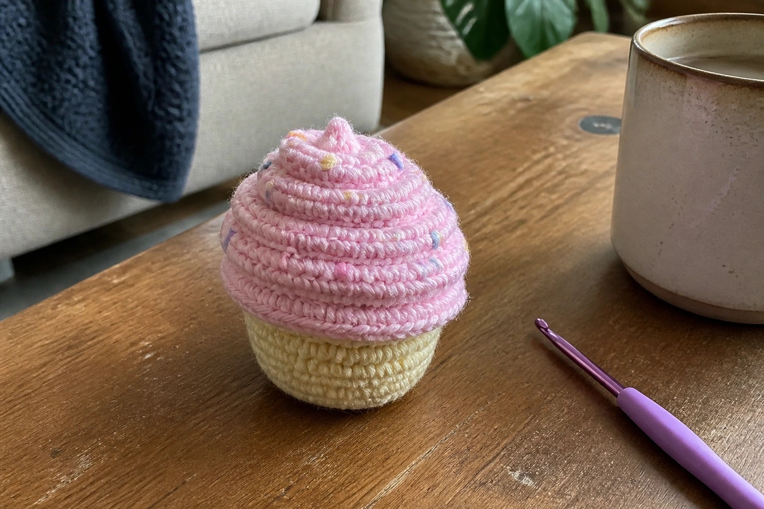 Pink crocheted cupcake on wooden table beside purple hook and mug