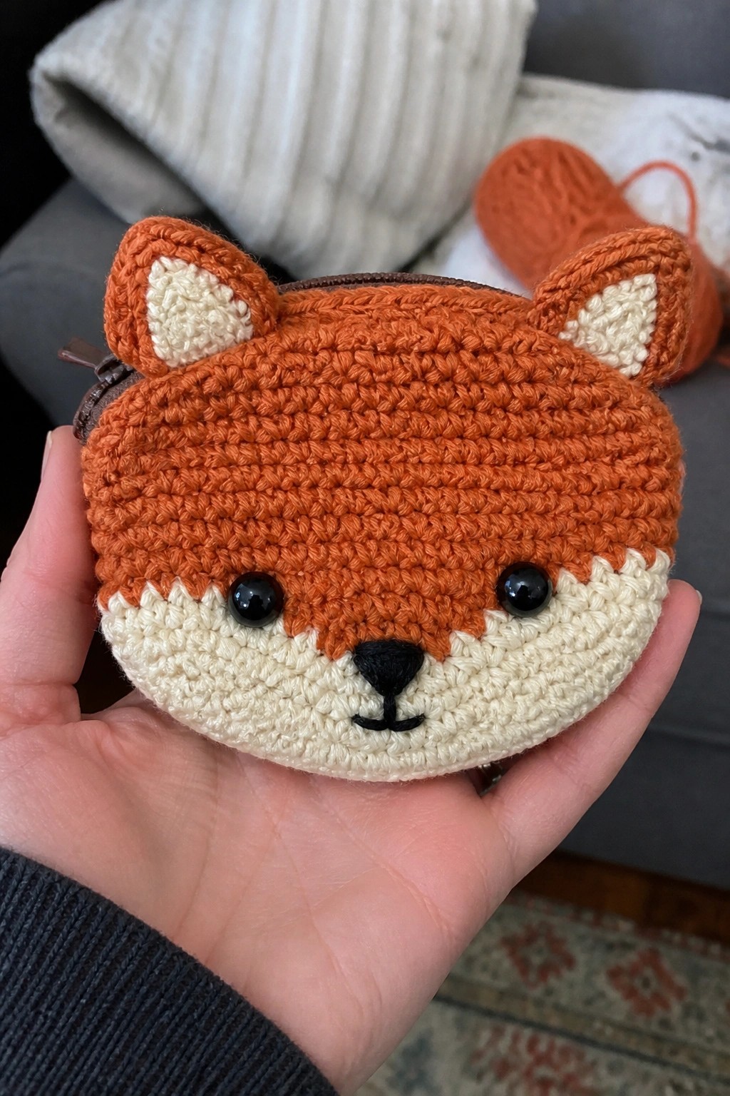 A hand holding a crocheted orange and cream fox face pouch with black eyes and nose and a small zipper on the left side.