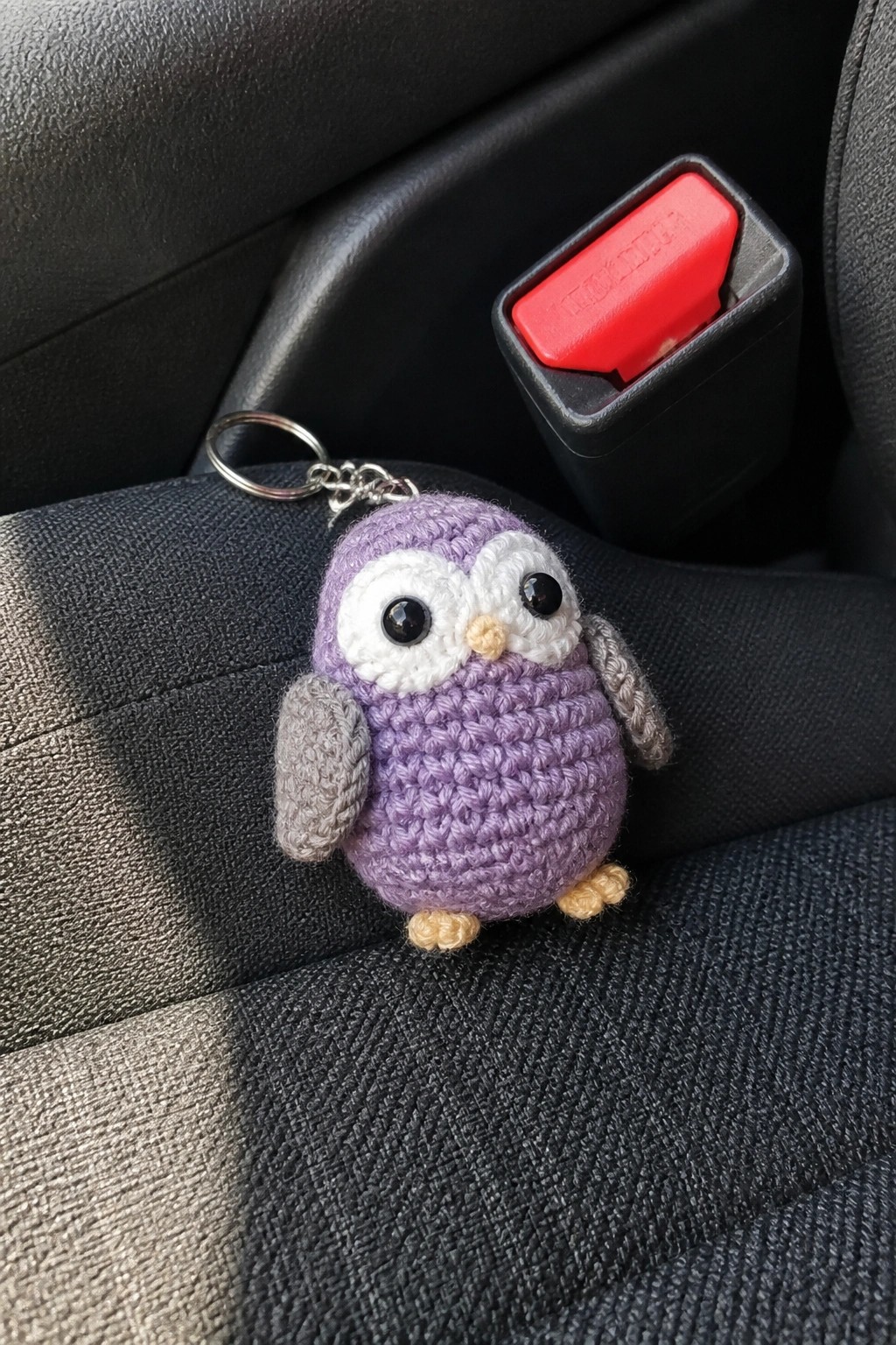 Purple crocheted owl keychain on car seat beside red seatbelt buckle.