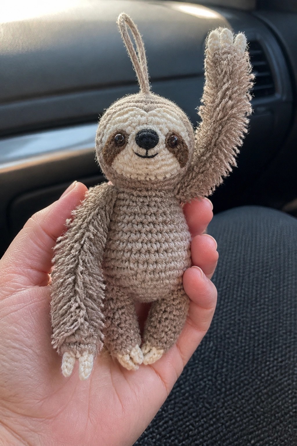 Hand holding crocheted sloth with one arm raised in a car