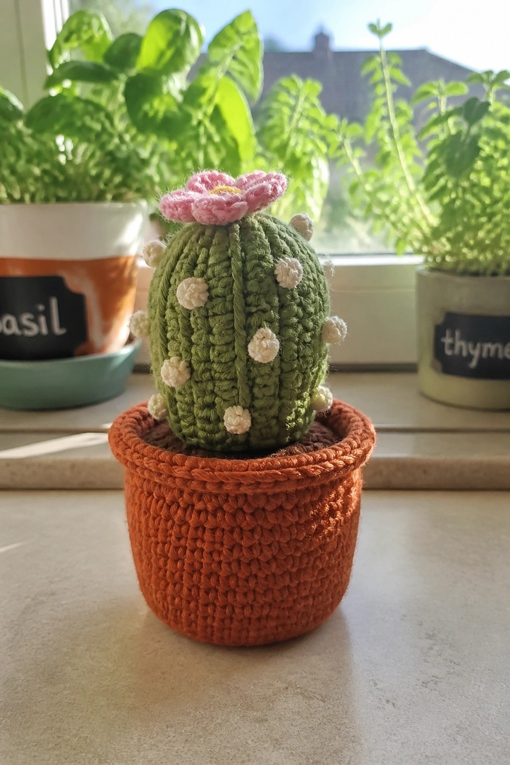 Crocheted green cactus with pink flower in orange pot on windowsill