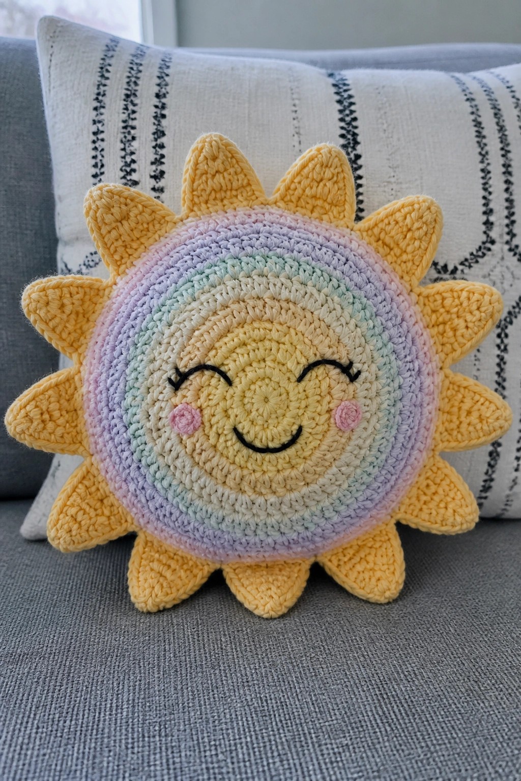 Crocheted sun plush with rainbow center, smiling face, and yellow rays on gray couch.
