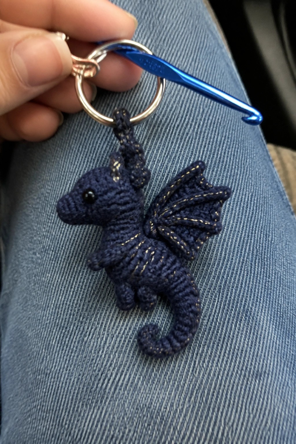 Hand holds crocheted navy dragon keychain with gold accents and blue hook over jeans