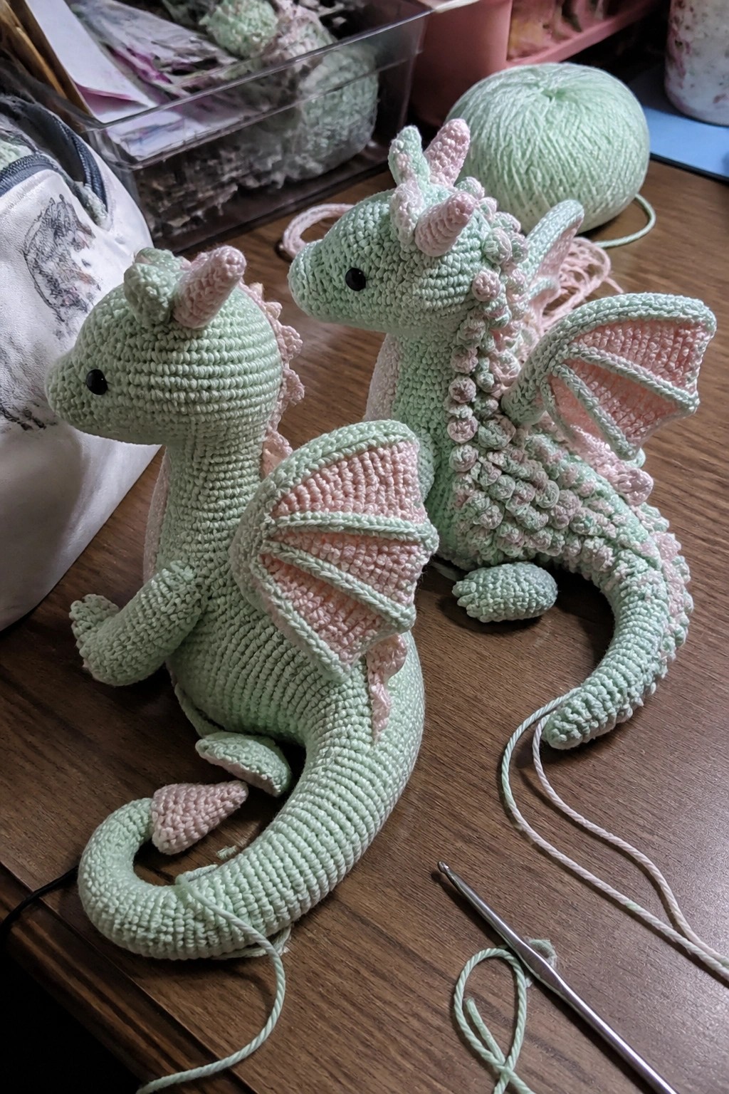 Two mint green crocheted dragons with pink wings on wooden table with yarn and hook