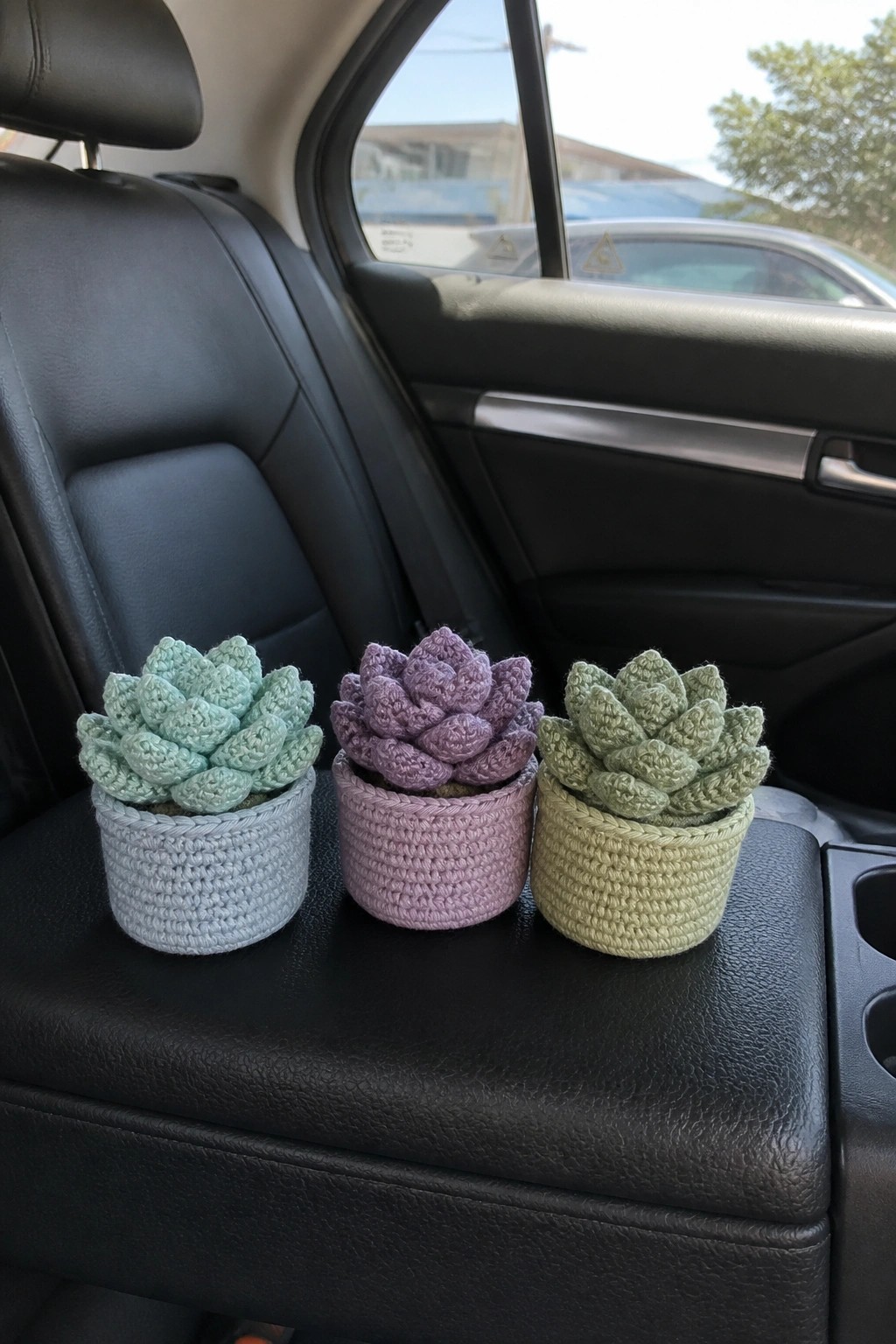 Three colorful crocheted succulents in pots on a black leather car seat.