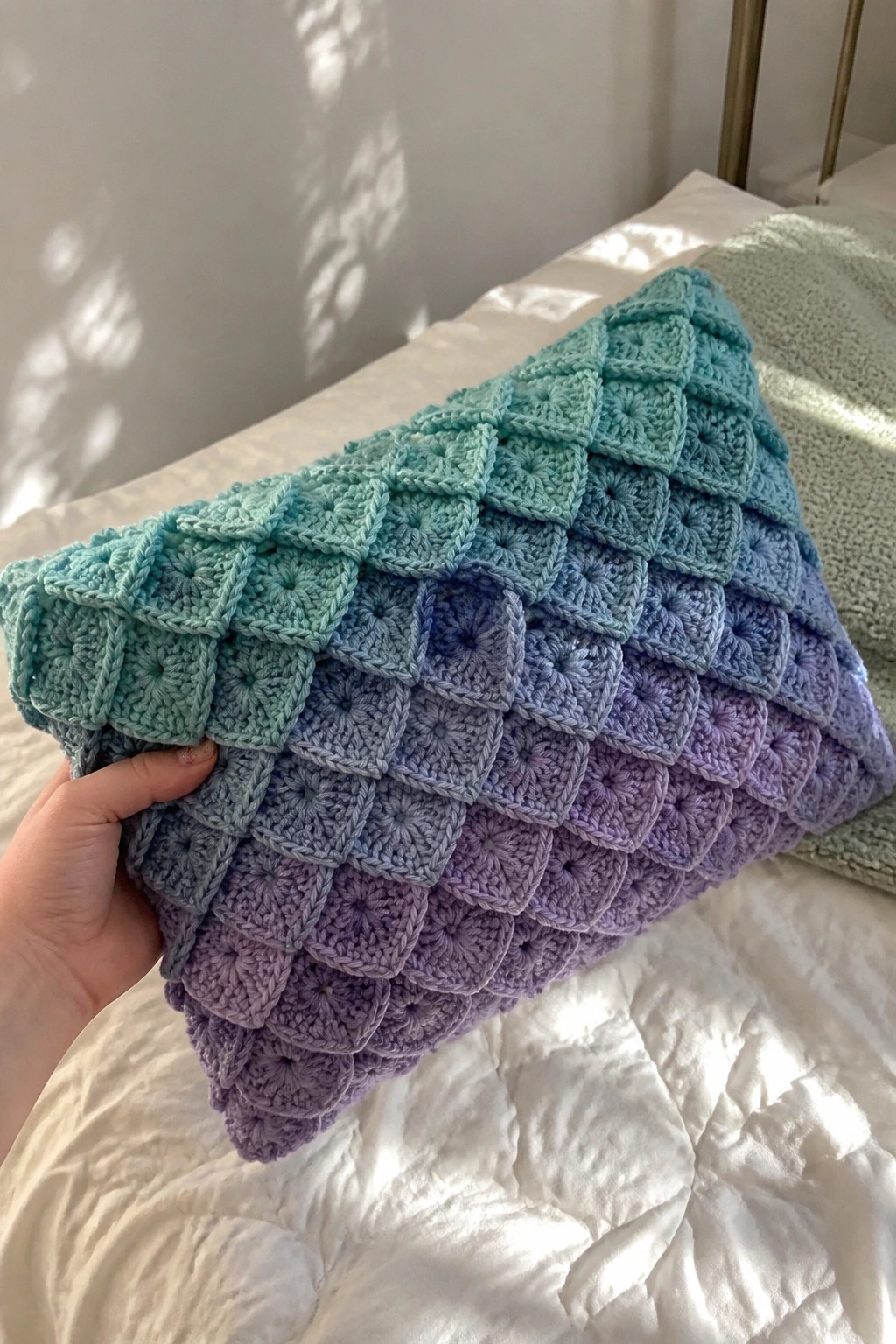 Hand holding gradient teal-to-purple granny square crochet blanket on white bed.