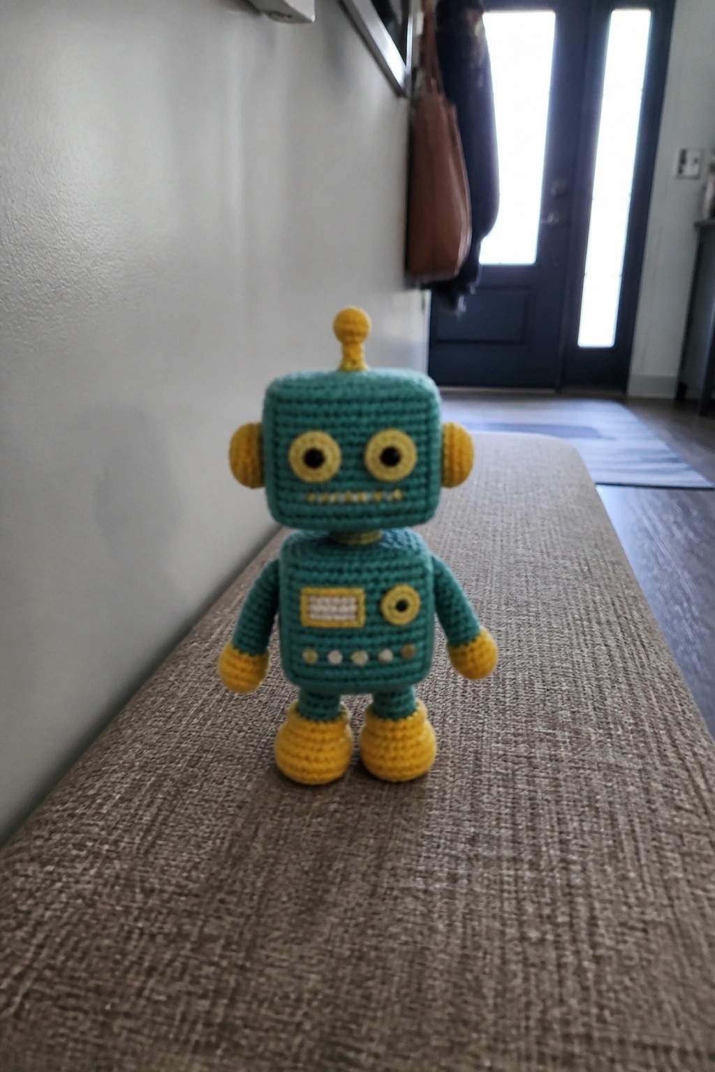 Crocheted teal robot toy with yellow accents stands on woven fabric surface.