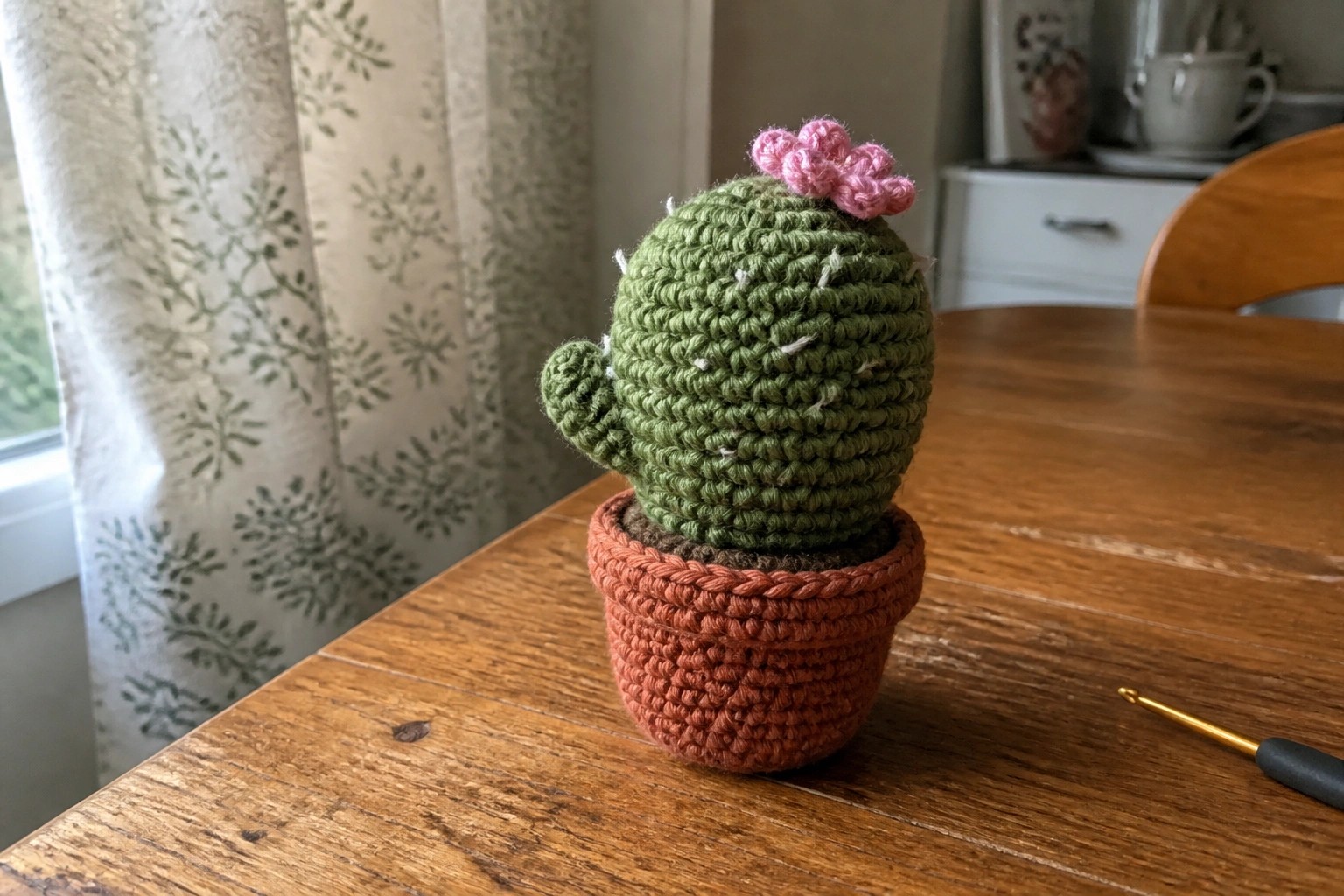 Green crocheted cactus with pink flower in orange pot beside gold hook on table