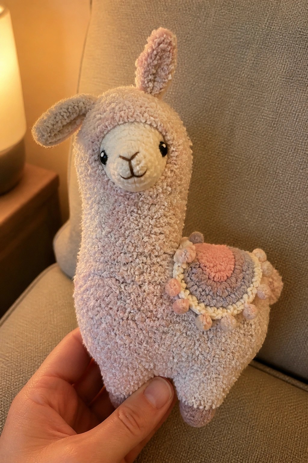 Hand holding fluffy light-pink llama plush with pastel pom-pom saddle.