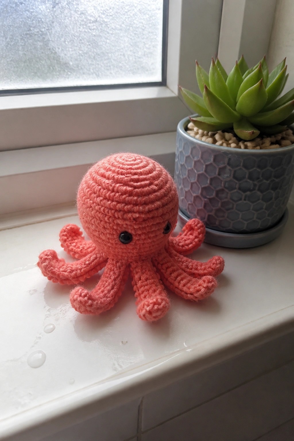 A small pink crocheted octopus sits on a white windowsill next to a potted green succulent.