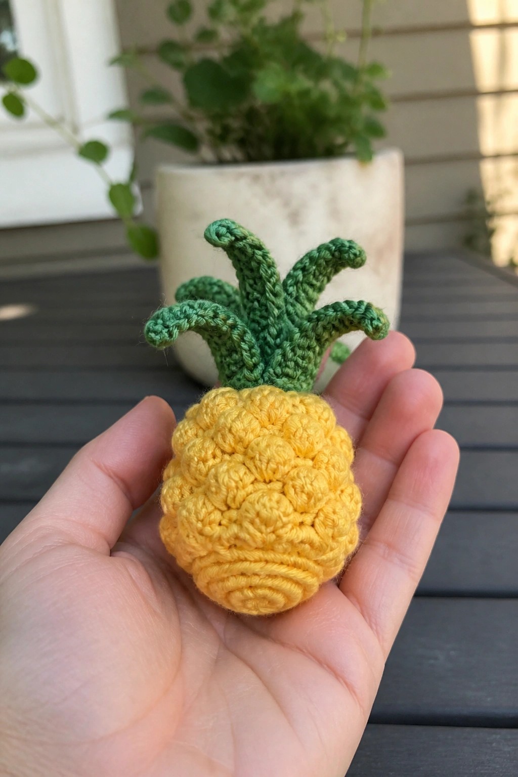 Hand holding yellow crocheted pineapple with green yarn leaves, potted plant behind.