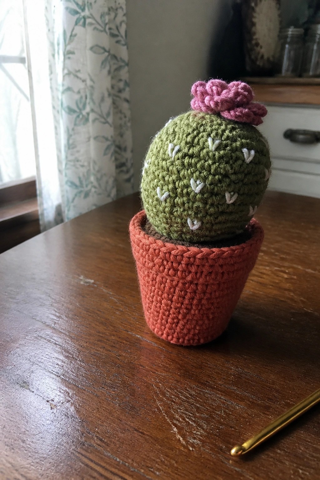 Green crocheted cactus with pink flower in orange pot beside gold hook on table