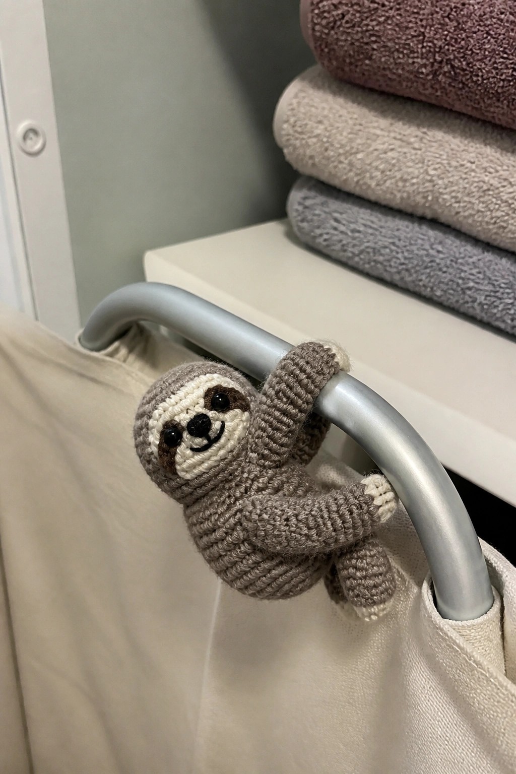 Crocheted sloth toy clinging to gray metal bar near stacked towels.