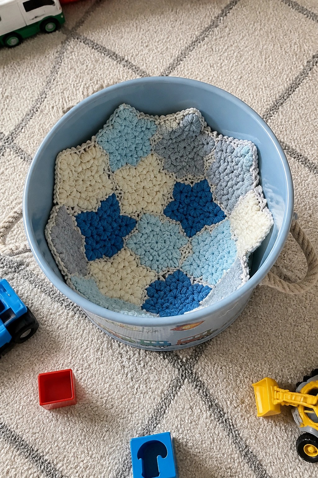 Blue bucket with colorful crocheted blanket and toy trucks on carpeted floor