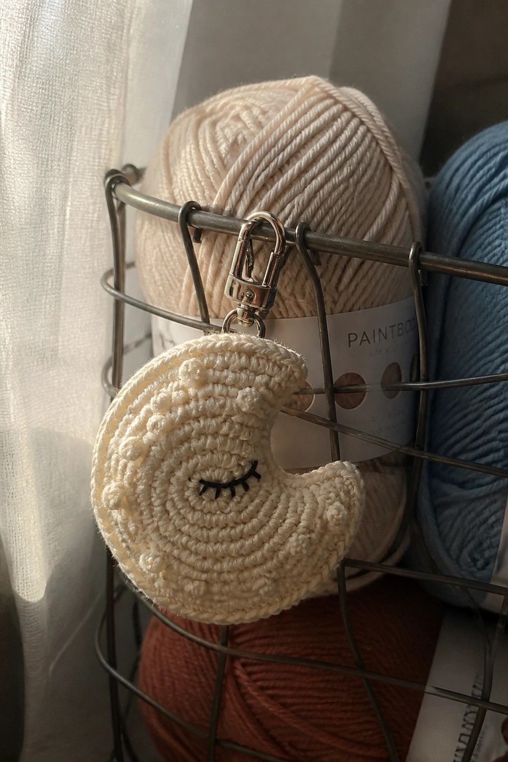 Crocheted crescent moon keychain with sleeping face on wire basket of yarn skeins