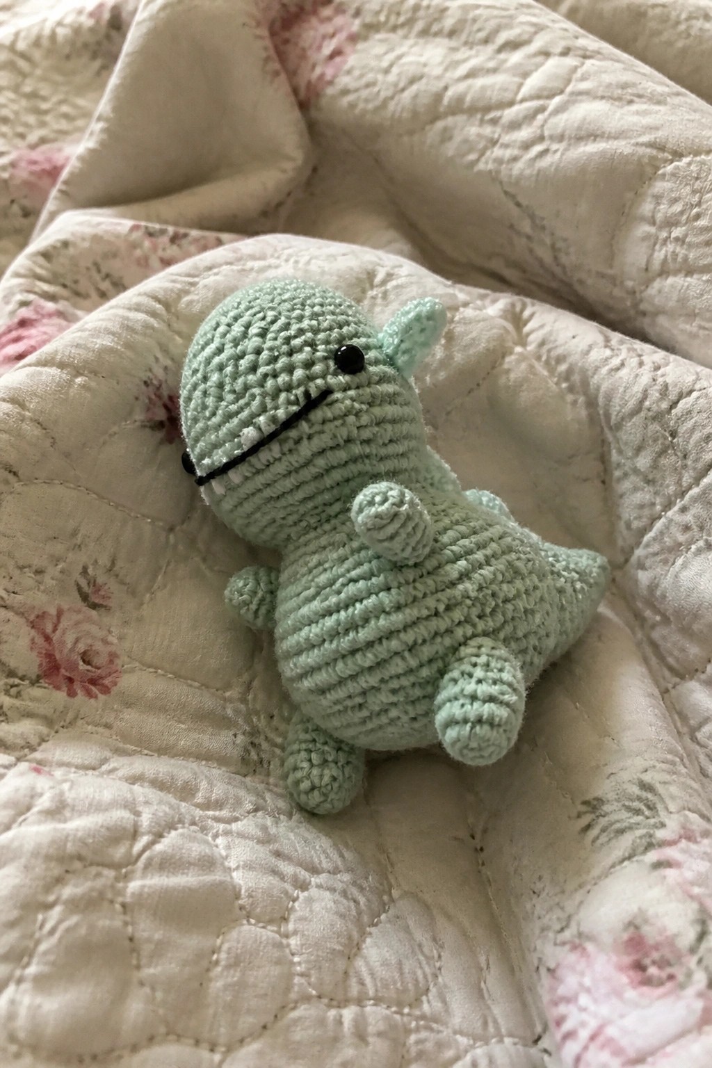 Mint green crocheted dinosaur toy on white floral quilted blanket.