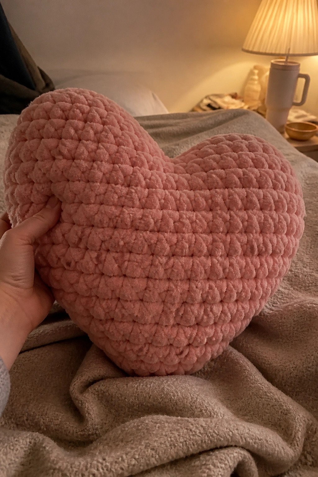 Hand holding large pink textured heart pillow on beige bedspread