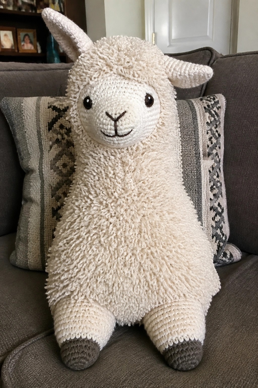 White crocheted llama plush toy seated on gray couch between patterned pillows