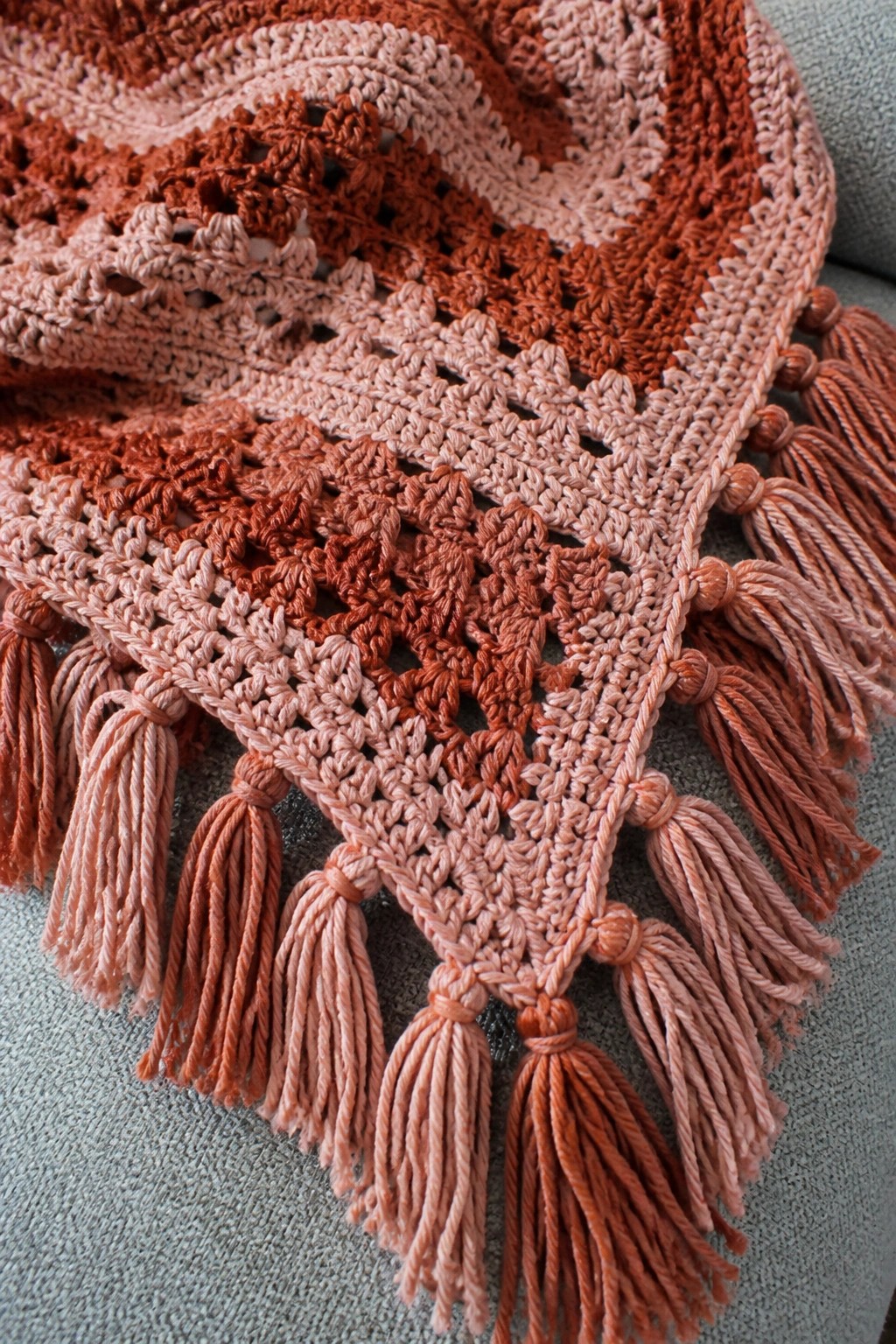 A crocheted blanket in pink and rust tones with lace motifs and tassels along the edges.