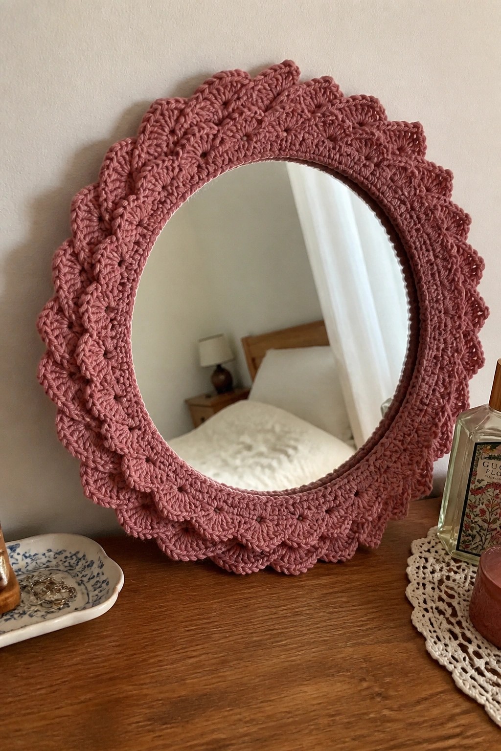 Pink crocheted round mirror with ruffled edge on wooden table beside perfume and candle