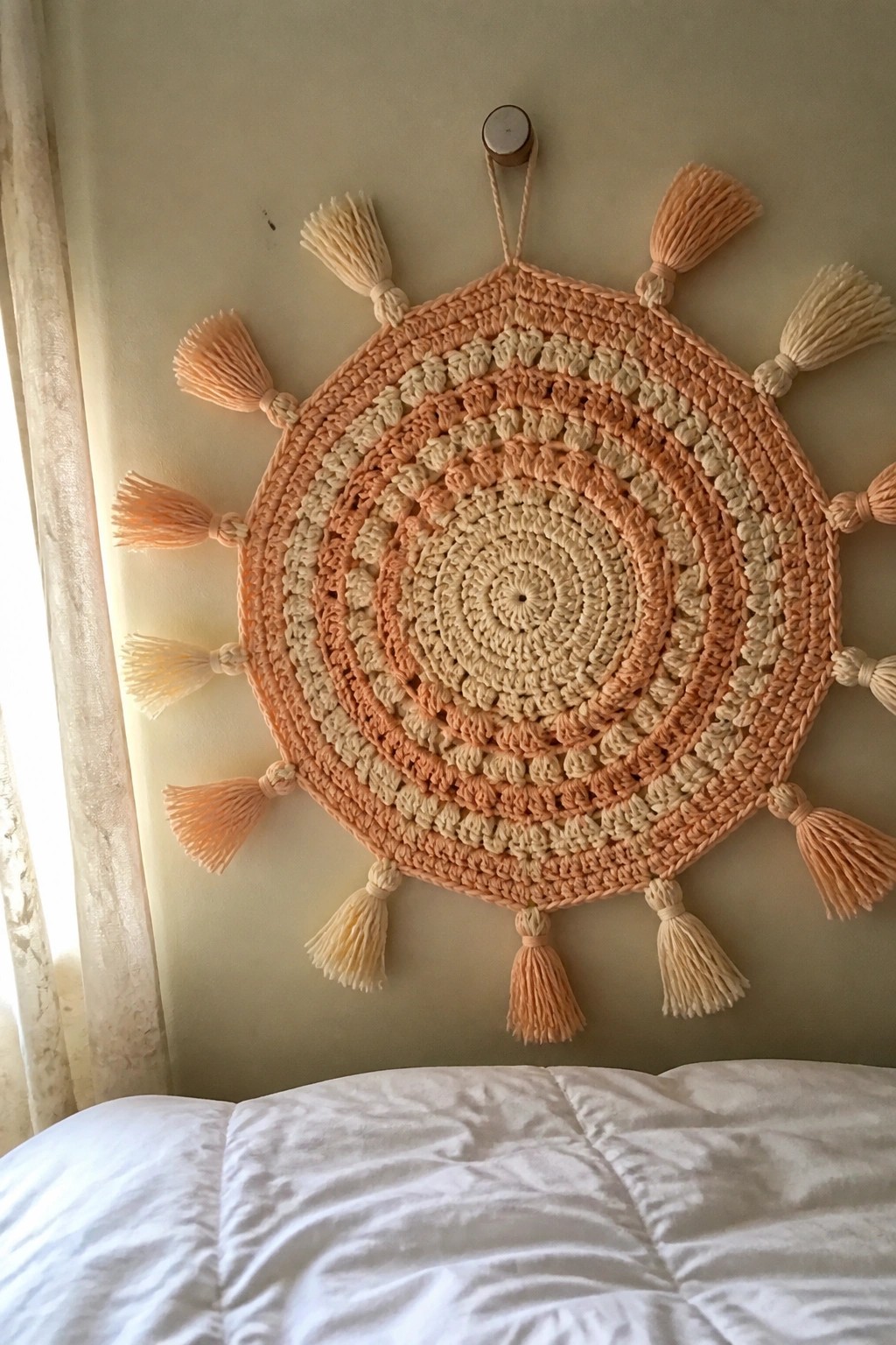 Peach and cream crocheted circular wall hanging with tassels above white bed.
