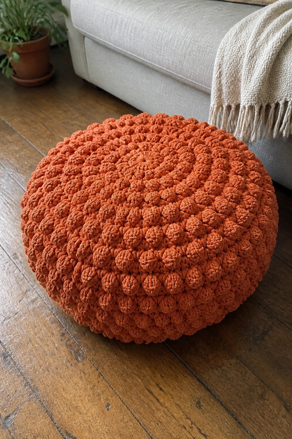 Orange crocheted pouf on wooden floor beside beige sofa with potted plant.