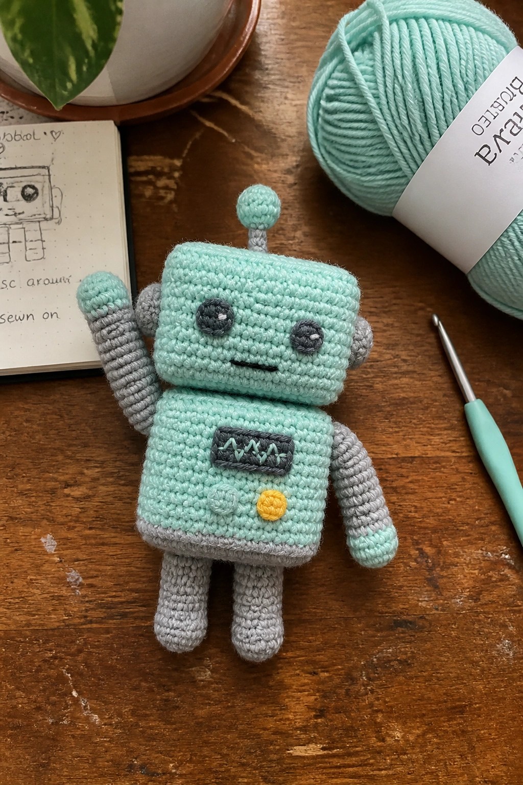 A crocheted mint green and gray robot amigurumi on a wooden table next to a sketchbook, yarn ball, and crochet hook.