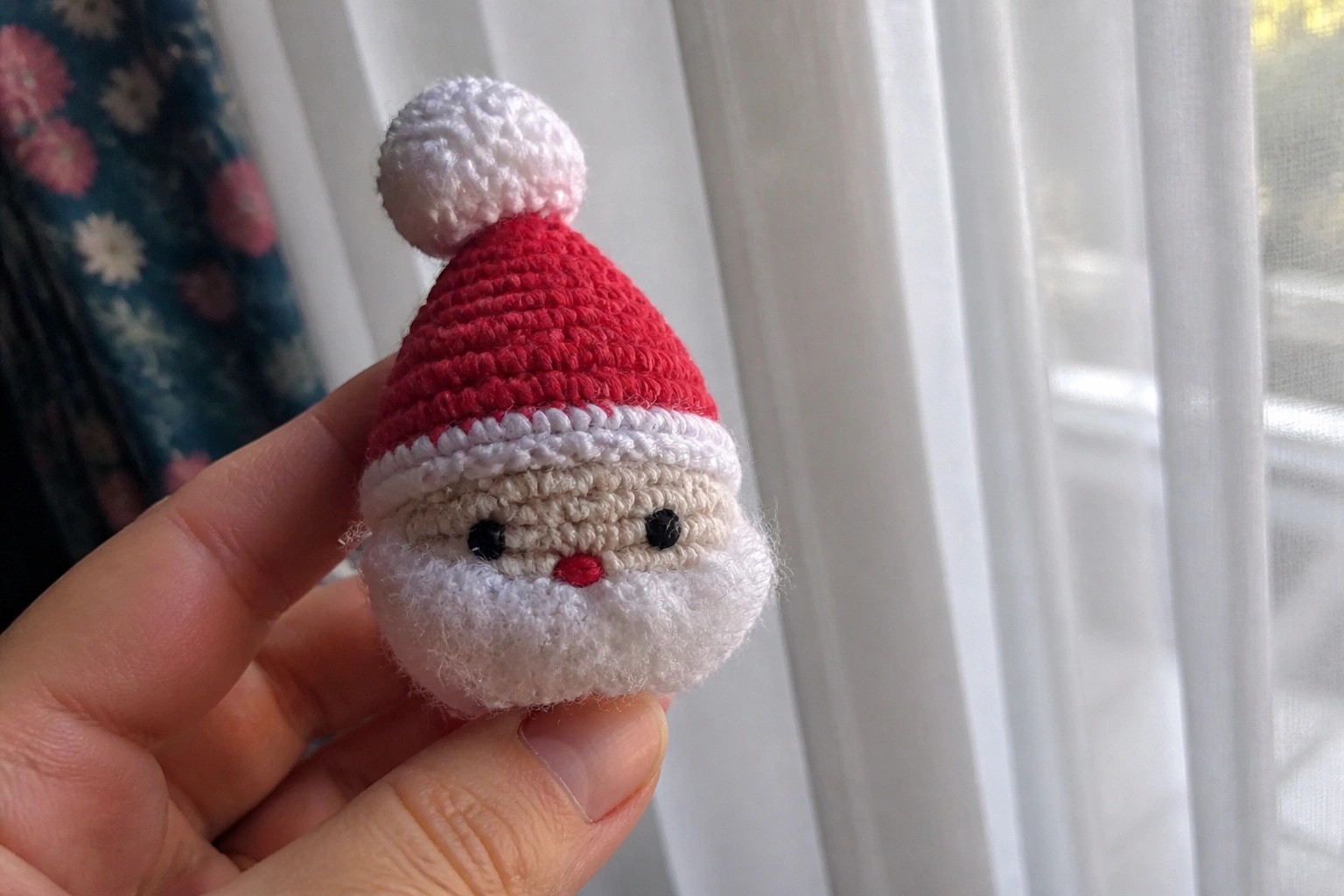 Hand holding miniature crocheted Santa with red hat, white beard, black eyes near window